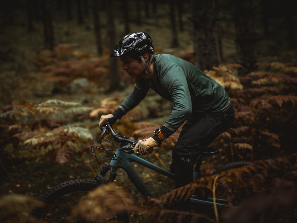 A mountain biker wearing a black helmet and a dark green long-sleeve jersey rides through a dense forest of ferns and trees. The bike frame is a muted teal blue.